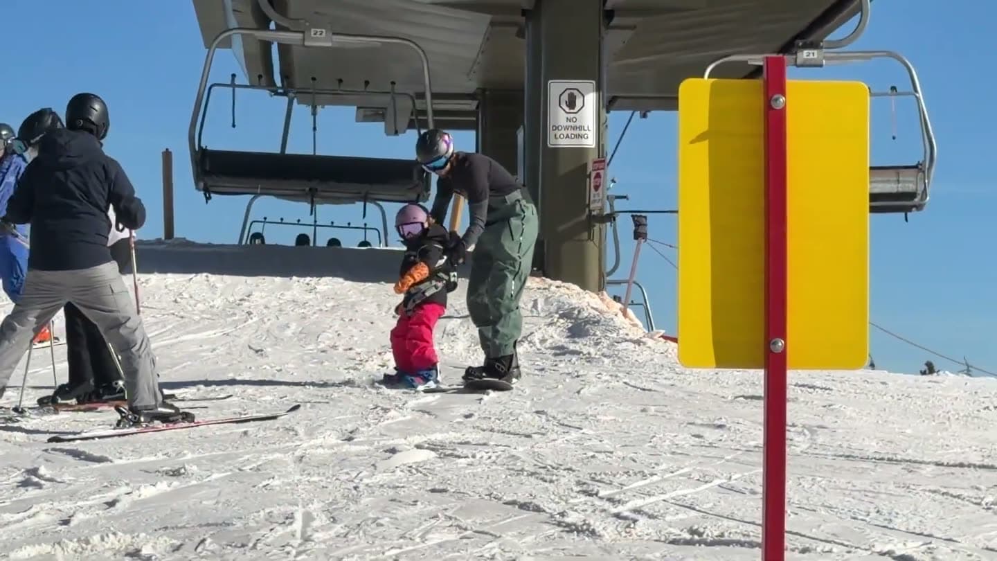 Parent helping a toddler snowboarder ride off the chairlift at the top