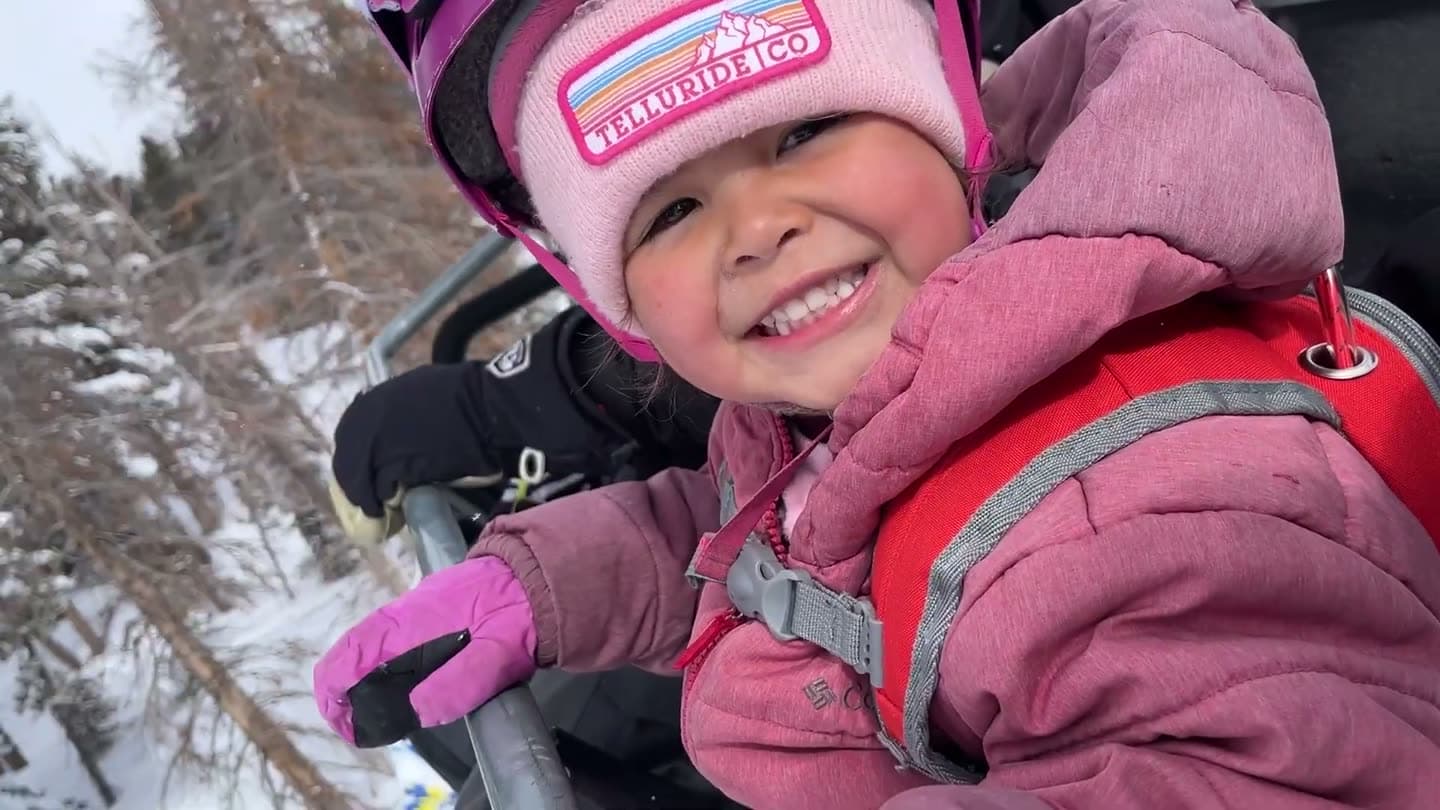 Smiling toddler in a helmet and harness backpack riding a chairlift