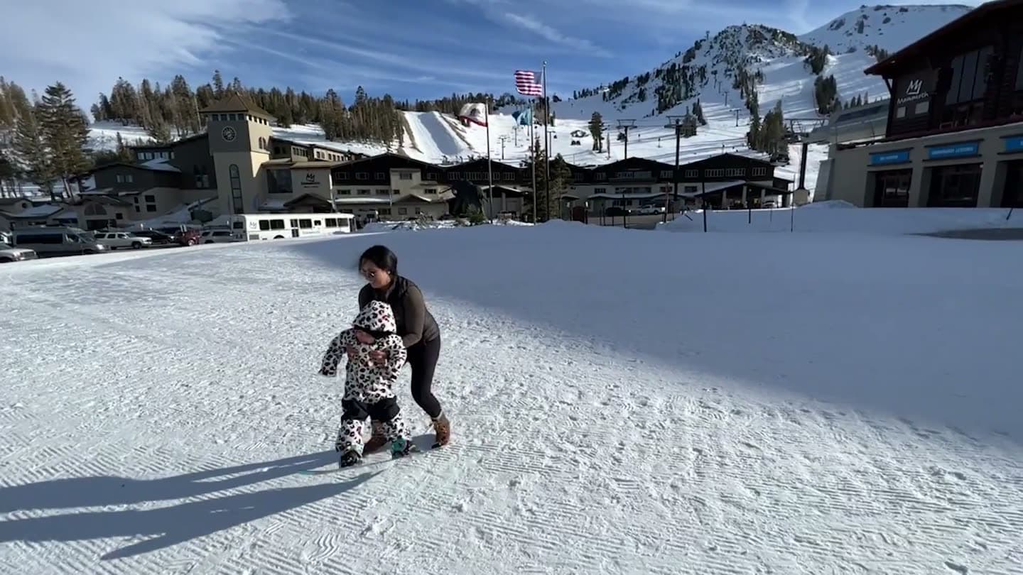 Parent pulling a toddler on a snowboard across flat snow near the base lodge