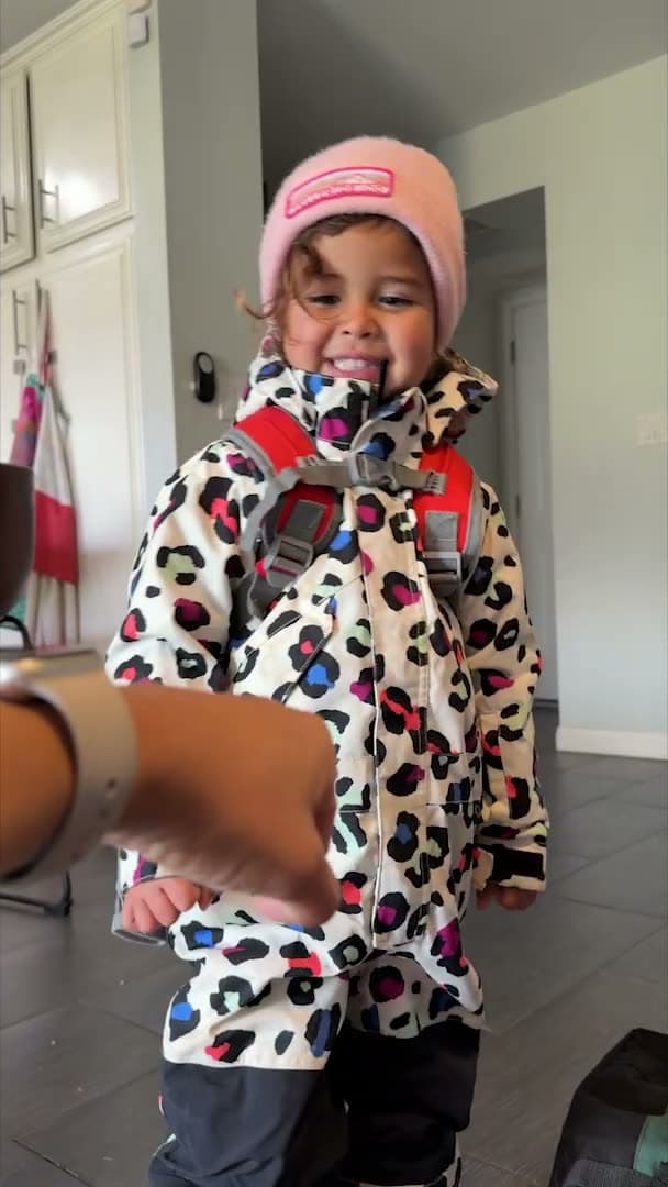 Toddler wearing her snowsuit and harness backpack at home before a snowboarding trip