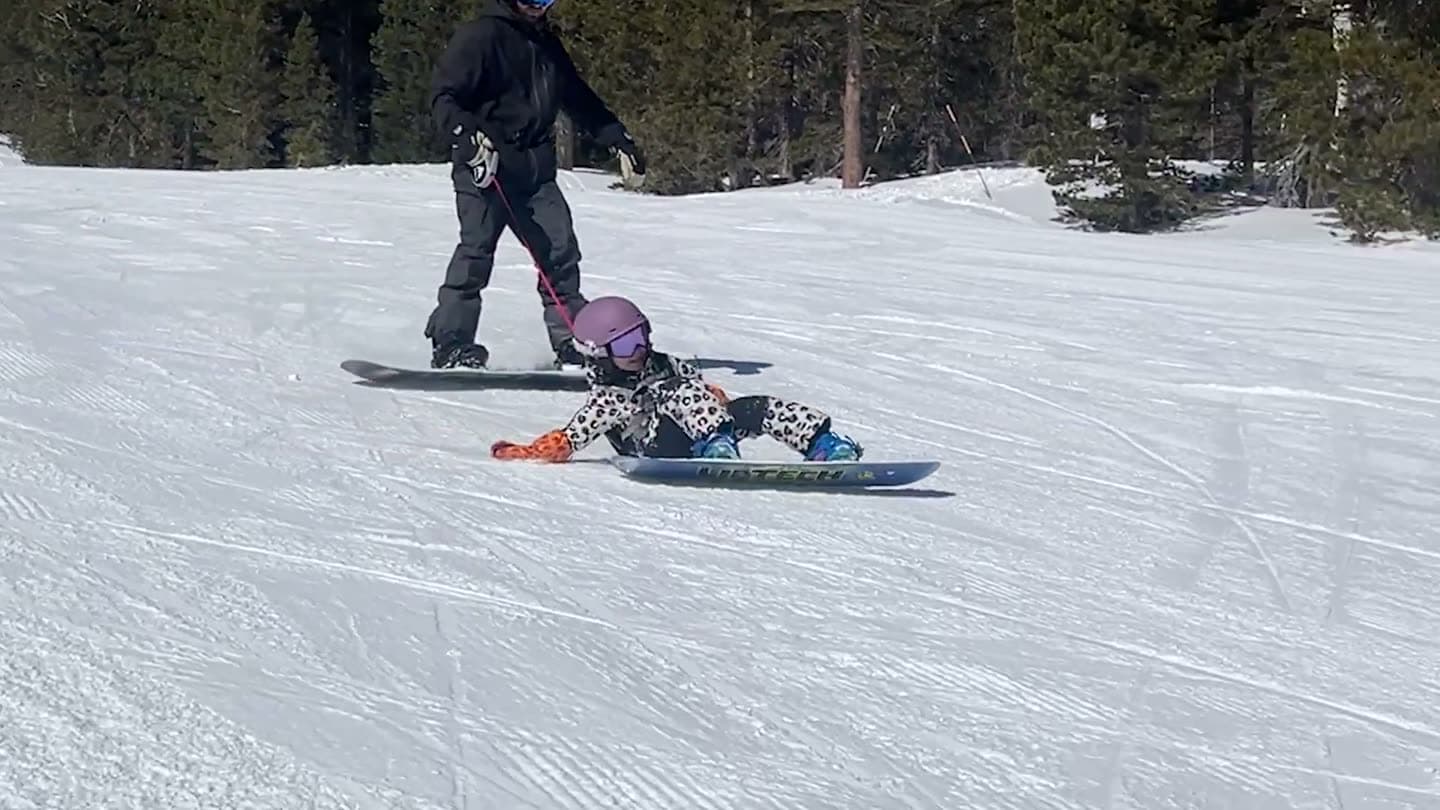 Toddler snowboarder sitting down after a fall while a parent holds her harness leash