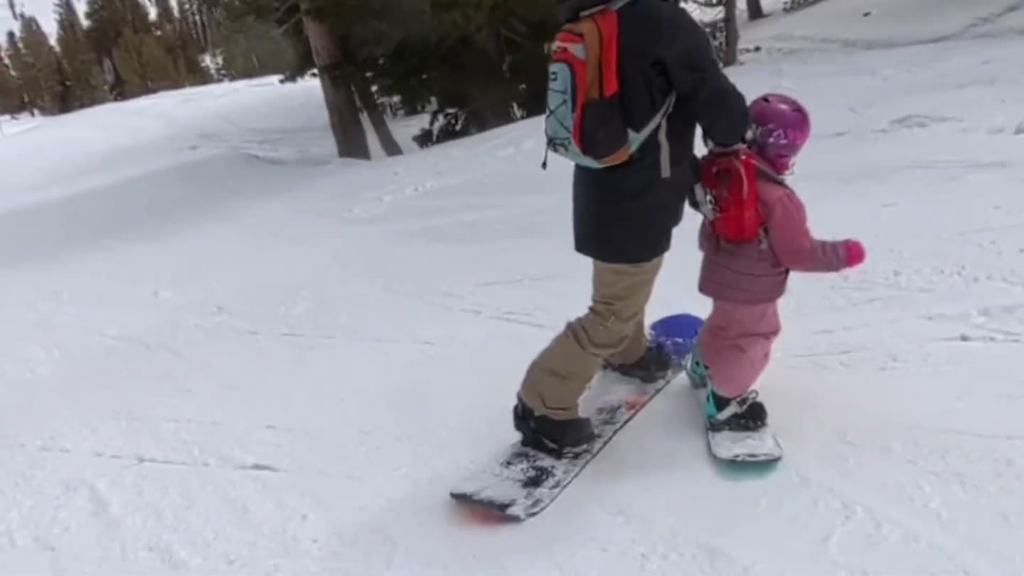 Parent snowboarding behind a toddler, holding the handle of her harness backpack