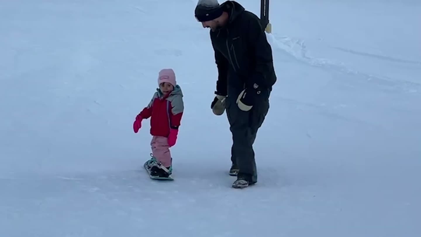 Dad walking alongside a toddler as she glides down a gentle slope on her snowboard